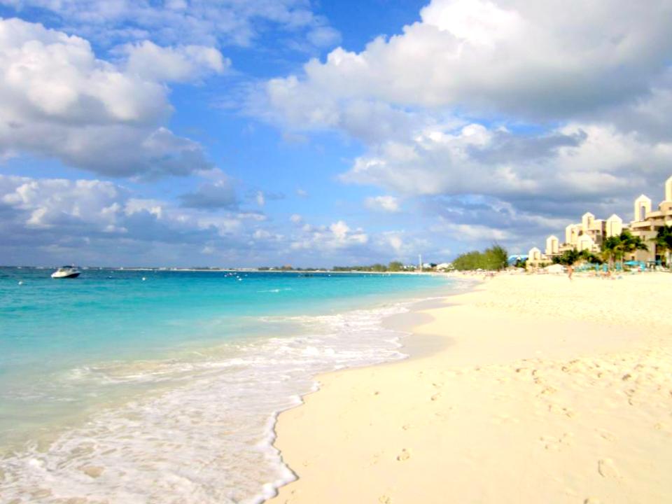 A picturesque beach scene with soft, white sand and clear, turquoise water. The sky is partly cloudy with patches of blue. There are buildings and palm trees along the right side of the beach, and a boat is visible in the water on the left side. The beach appears calm and inviting.