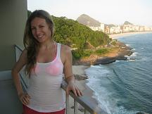 A woman is standing on a balcony overlooking a scenic coastal view. She is smiling and wearing a white tank top and red shorts. In the background, there are lush green hills, a beach, and buildings along the coastline. The ocean is visible to the right, with waves crashing against the shore.