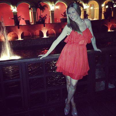 A woman in a red dress is posing and smiling while leaning against a wooden railing. Behind her, there is a water fountain and an illuminated building with arches and potted plants. The scene appears to be taken at night, with warm lighting creating a cozy atmosphere.