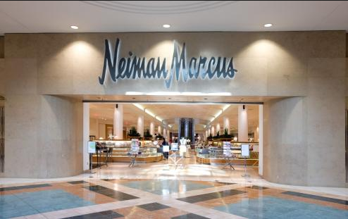 The image shows the entrance to a Neiman Marcus store. The store's name is prominently displayed above the entrance in large, stylish letters. The entrance is wide and open, with a view into the well-lit interior of the store, which features various displays and merchandise. The floor in front of the entrance has a patterned design with different shades of tiles.