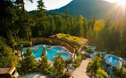 The image shows an outdoor spa resort nestled in a forested area with a mountainous backdrop. There are multiple pools of varying sizes and shapes, surrounded by lush greenery and tall trees. A pathway winds through the area, connecting the pools and leading to a building with a green roof. The sun is setting or rising, casting a warm glow over the scene.