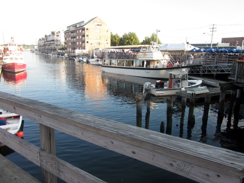 A waterfront scene featuring a wooden boardwalk in the foreground. Several boats are docked along the water, including a large tour boat with passengers on the upper deck. The water reflects the buildings and boats, creating a picturesque view. In the background, there are multi-story buildings and trees, with a clear sky above. The area appears to be a lively and popular spot, possibly a marina or harbor.