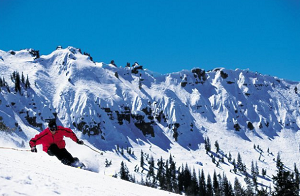 A person wearing a red jacket is skiing down a snowy slope with a backdrop of rugged, snow-covered mountains and a clear blue sky. Pine trees are visible at the base of the slope.