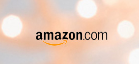 The image shows the Amazon.com logo, which consists of the word "amazon" in black lowercase letters followed by ".com" in a smaller font. There is a curved arrow underneath the word "amazon" that starts at the letter "a" and points to the letter "z," symbolizing that Amazon offers everything from A to Z. The background is blurred with soft, warm lights.