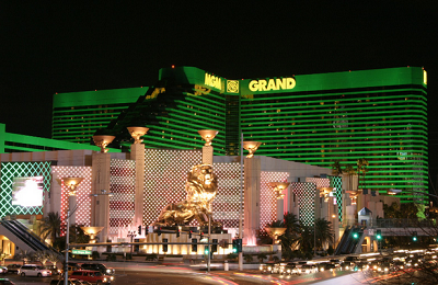 The image shows the MGM Grand hotel and casino in Las Vegas at night. The building is illuminated with green lights, and the large golden lion statue, which is a prominent feature of the MGM Grand, is visible in front of the hotel. The surrounding area is lit up with various lights, and there is some traffic visible on the street in front of the hotel.