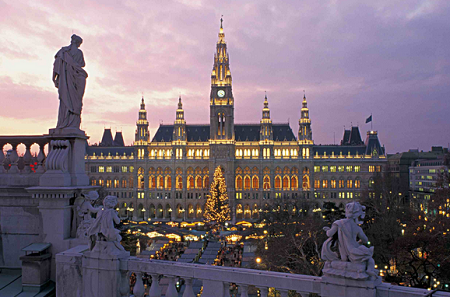 The image shows a grand, illuminated building with a tall central tower and multiple smaller spires, likely a historic or government building, set against a twilight sky. In front of the building, there is a large, decorated Christmas tree surrounded by festive lights and market stalls. Statues and sculptures are visible in the foreground, adding to the architectural beauty of the scene. The overall atmosphere is festive and elegant.