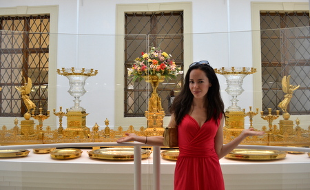 A woman in a red dress is standing in front of a display of ornate golden artifacts, including candlesticks, trays, and a floral arrangement. The background features windows with decorative grilles. The woman has her arms slightly raised and is smiling at the camera.