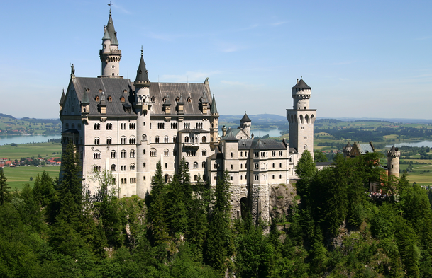 The image shows a large, picturesque castle with multiple towers and spires, situated on a forested hill. The castle has a fairytale-like appearance with its white walls and dark roofs. In the background, there is a scenic landscape featuring rolling hills, a lake, and a clear blue sky.