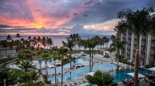 The image shows a luxurious resort with multiple swimming pools surrounded by palm trees. The resort is located by the ocean, and the sun is setting, casting a beautiful array of colors across the sky, including shades of pink, purple, and orange. The scene is tranquil and picturesque, with lounge chairs arranged around the pools and the ocean visible in the background.