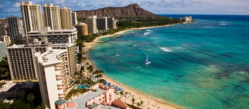 A coastal cityscape featuring a beach with turquoise waters and numerous people swimming and sunbathing. High-rise buildings and hotels line the shore, with a prominent mountain in the background. The sky is partly cloudy, and the overall scene is vibrant and tropical.