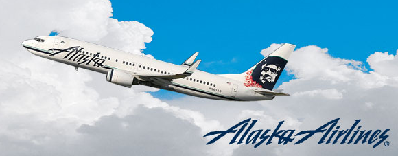 An Alaska Airlines airplane is flying against a backdrop of blue sky and white clouds. The aircraft features the Alaska Airlines logo and a stylized image of a person's face on its tail. The "Alaska Airlines" text logo is also visible in the bottom right corner of the image.